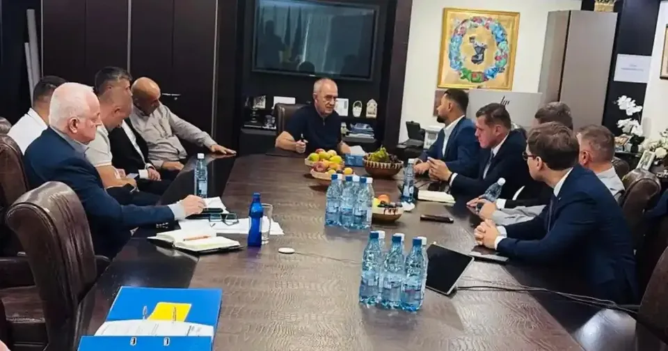 Mai mulți bărbați stau în jurul unei mese lungi, cu sticle de apă, pahare, documente și fructe, într-o încăpere cu mobilier închis la culoare.