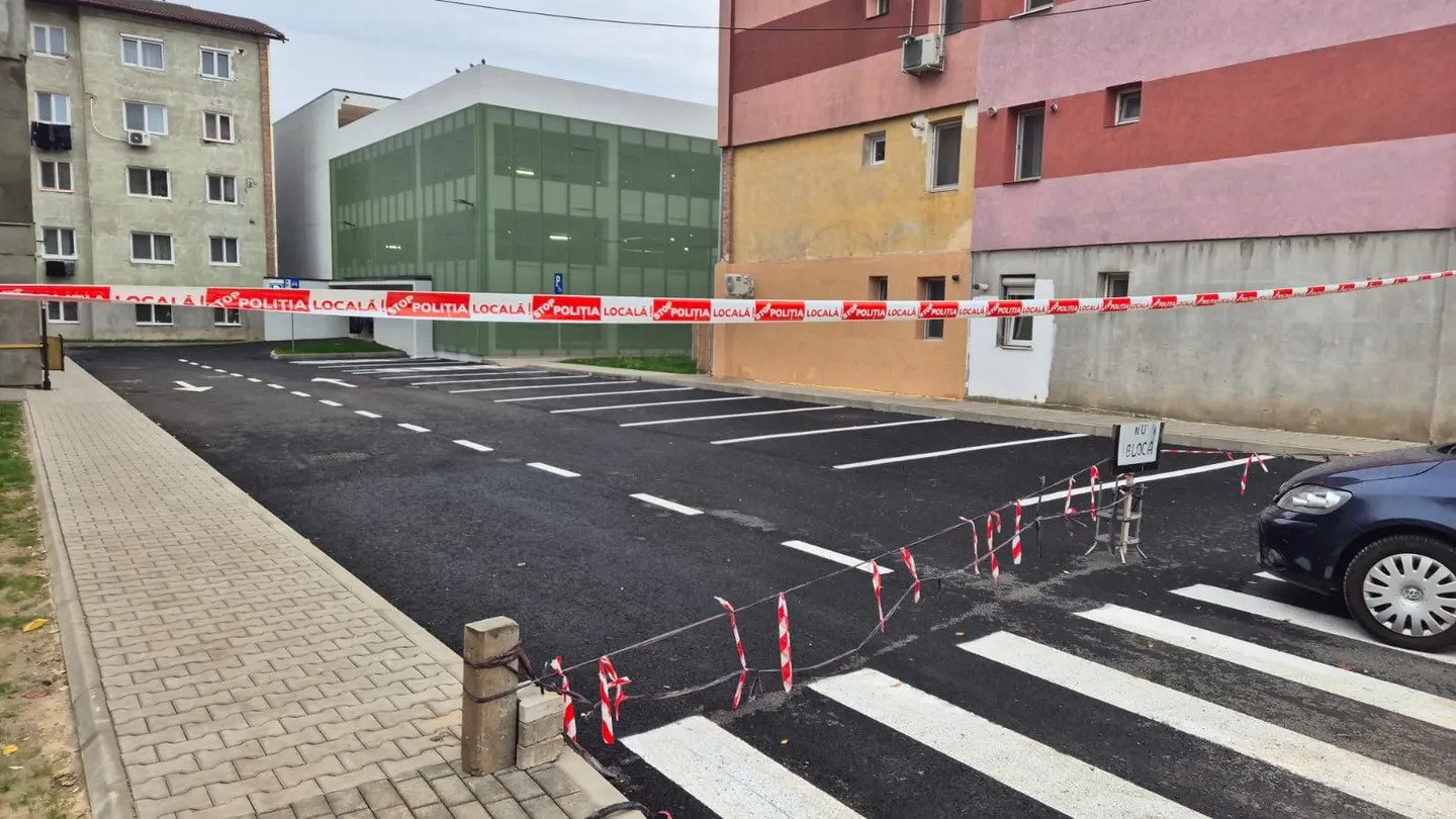 Benzi roșii și albe traversează carosabilul și delimitează o trecere de pietoni; în jur se văd clădiri, un trotuar și un autoturism.