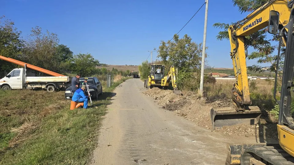 Pe un drum îngust se văd două utilaje galbene, pământ excavat, o autoutilitară albă, un autoturism închis la culoare și persoane lângă acostament.