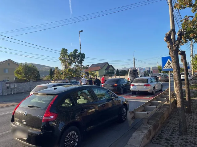 Un autoturism negru cu partea din spate avariată se află lângă o bordură; alte autoturisme și câteva persoane sunt vizibile pe șosea.