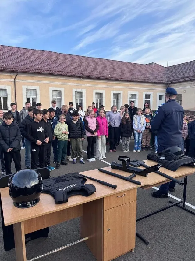 Un bărbat în uniformă stă în fața unui grup de copii, lângă o masă cu o cască, veste și alte echipamente negre.