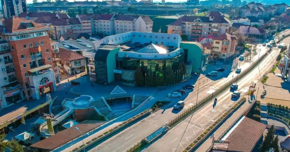 O clădire mare, cu fațadă curbată și acoperiș deschis la culoare, se află lângă o stradă cu câteva mașini; în jur sunt alte clădiri.