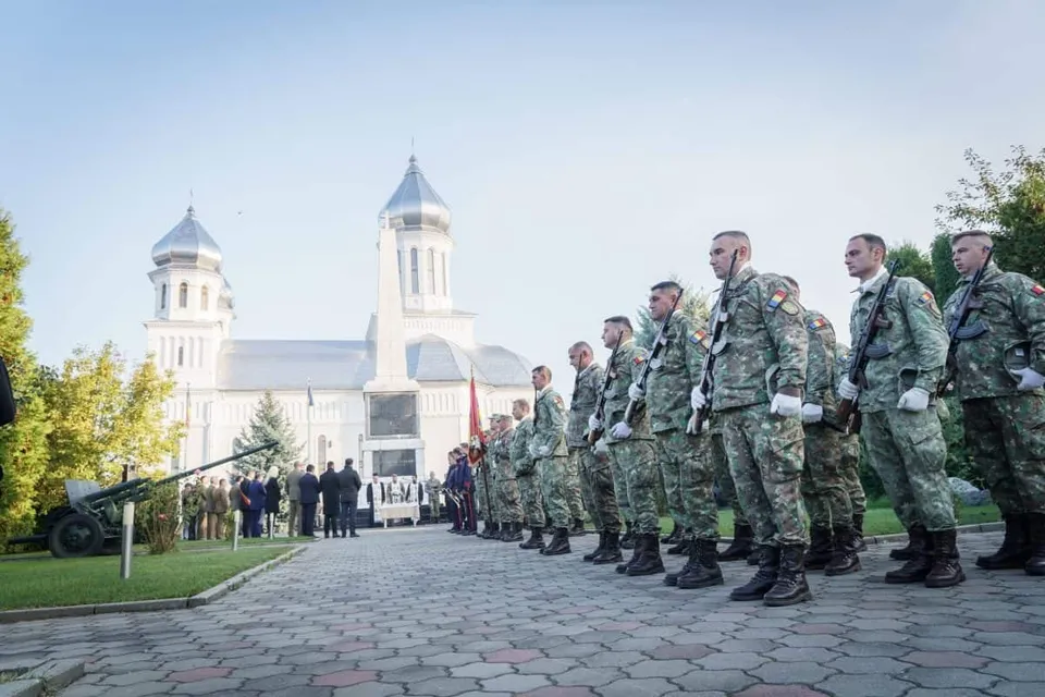 Militari în uniforme de camuflaj stau aliniați pe o alee pavată; în fundal se văd alte persoane, un tun și o clădire albă cu turnuri.
