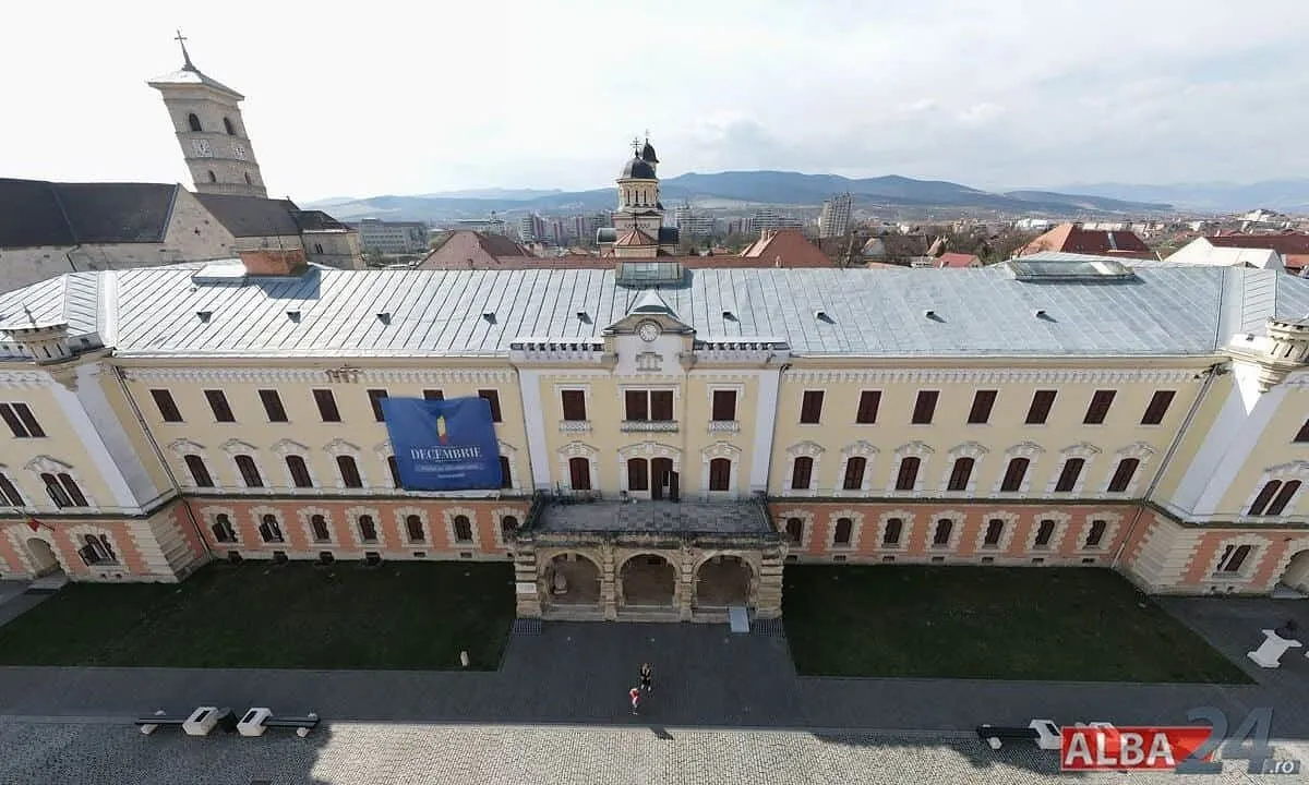 O clădire lungă, cu acoperiș deschis la culoare și intrare cu arcade, se află lângă o alee cu câteva mașini și persoane mici.