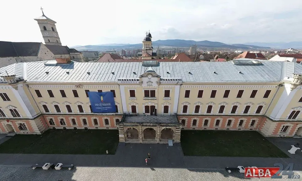O clădire lungă, cu acoperiș deschis la culoare și intrare cu arcade, se află lângă o alee cu câteva mașini și persoane mici.