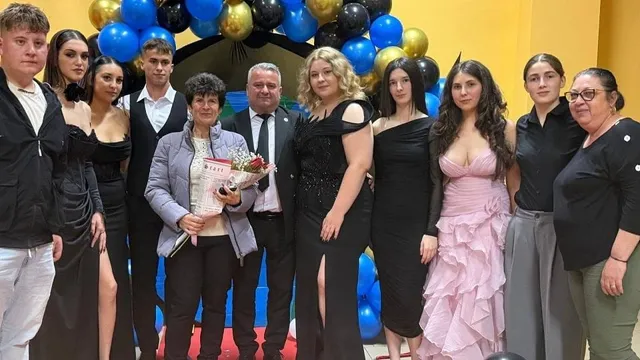 Un grup de persoane stă în fața unui arc de baloane albastre, negre și aurii; o femeie ține un document și un buchet.