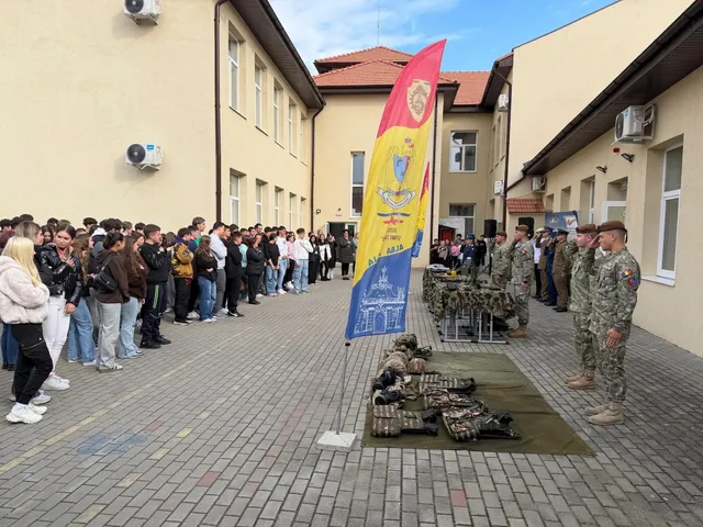 Un grup de tineri stă lângă două clădiri, în fața unor persoane în uniforme militare și a unor echipamente așezate pe covoare.