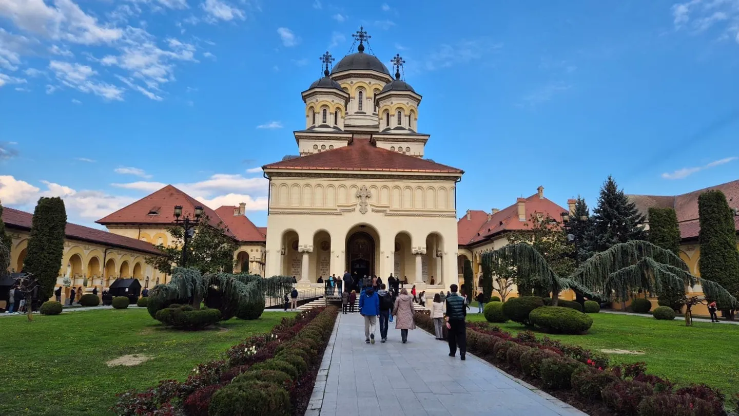 O clădire cu arcade și cupole se află în fundal, în spatele unei alei pavate, pe care merg mai multe persoane.