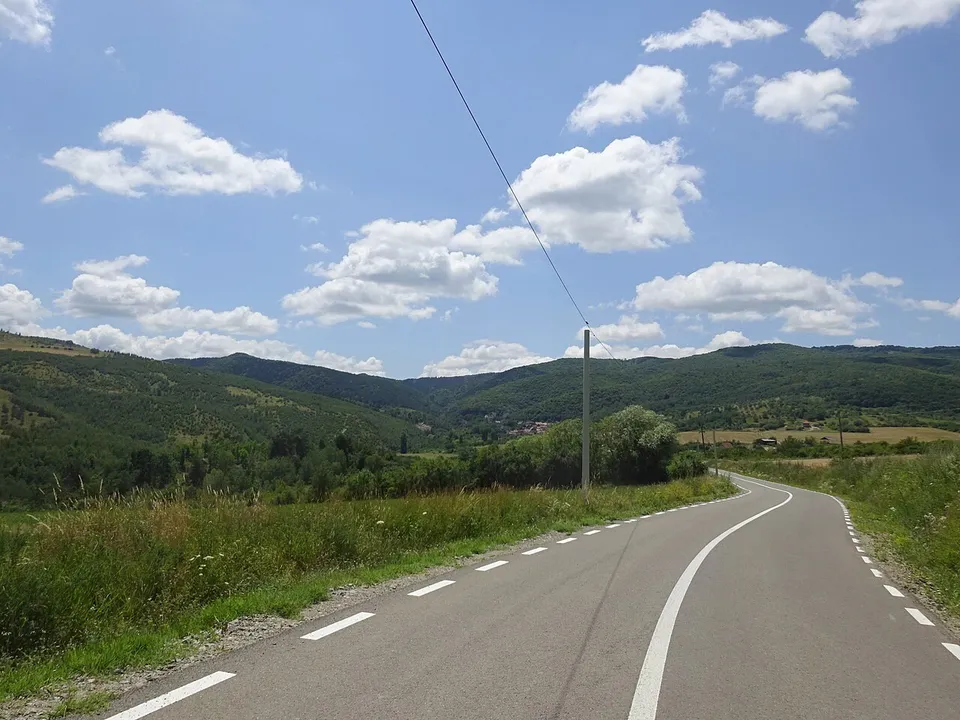 Un drum asfaltat cu marcaje curbe spre dreapta, mărginit de vegetație, cu dealuri verzi, un stâlp și nori pe cer.