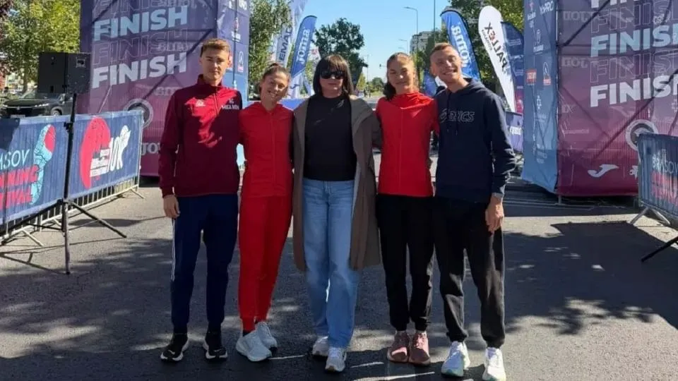 Cinci persoane stau aliniate între garduri metalice, în fața unor panouri cu inscripția „FINISH”, purtând haine sport roșii, negre și albastre.