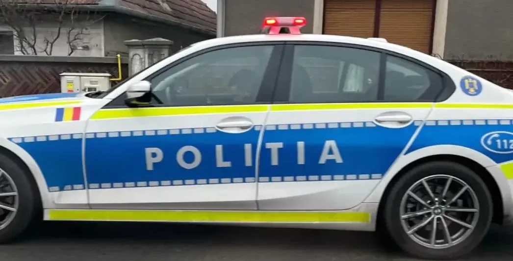 Un autoturism alb și albastru, inscripționat „POLIȚIA”, este văzut din lateral, cu o bară luminoasă pe plafon și clădiri în fundal.