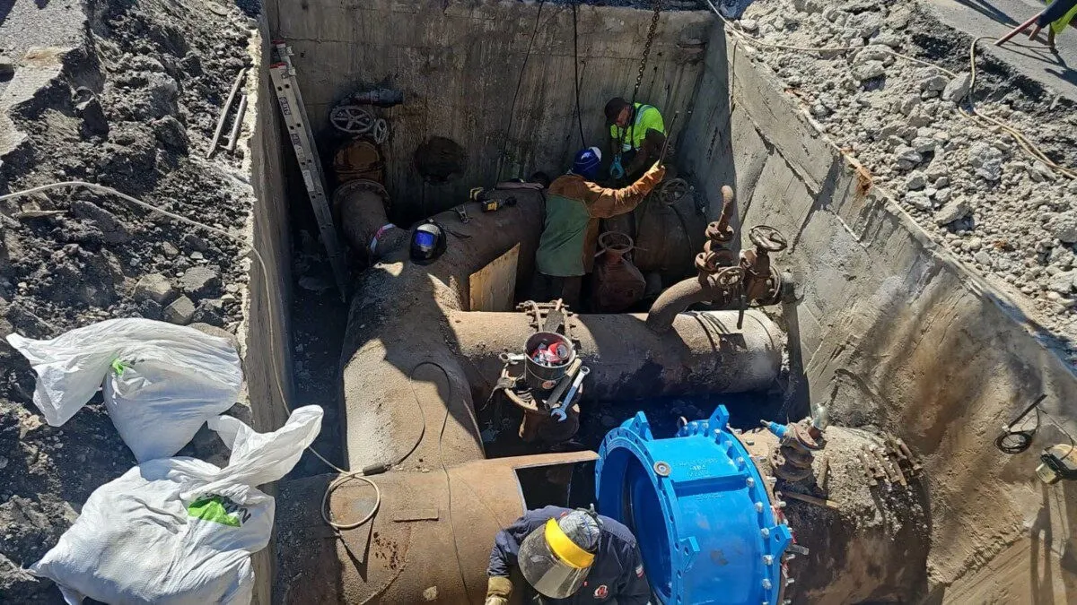 Într-o groapă cu pereți de beton, trei persoane cu căști lucrează lângă conducte groase, un robinet și o piesă albastră.