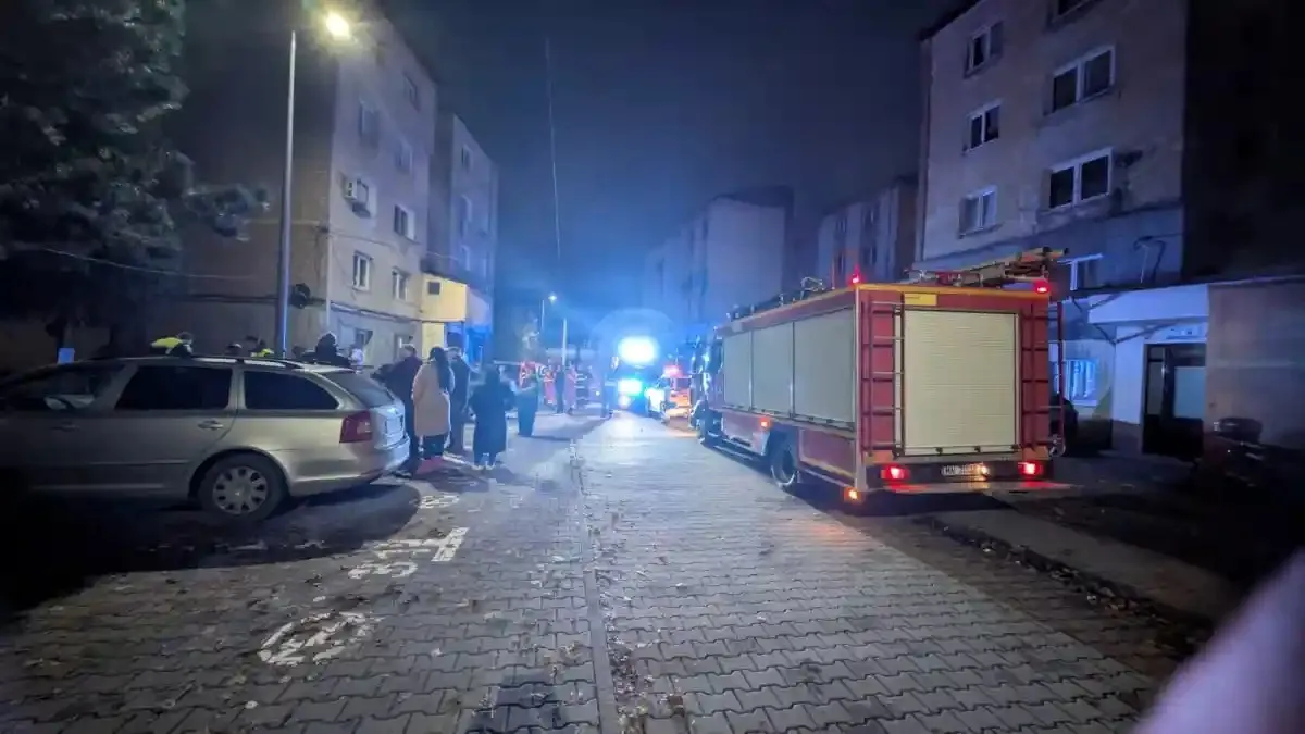 Pe o stradă nocturnă se văd o autospecială roșie, un autoturism argintiu, câteva persoane și lumini albastre între clădiri.