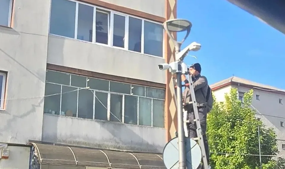 Un bărbat îmbrăcat în haine închise la culoare stă pe o scară lângă un stâlp cu camere video, în fața unei clădiri.