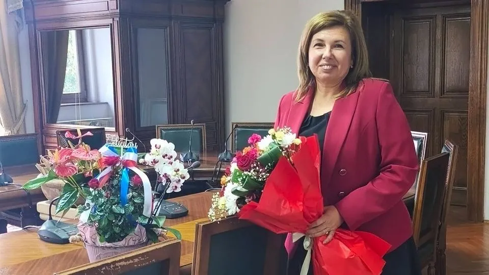 O femeie în sacou roșu ține un buchet de flori lângă o masă cu aranjamente florale, în fața unor dulapuri din lemn.