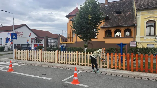 O persoană aplecată lângă un gard din șipci colorate ține mâinile pe gard; pe carosabil se văd două conuri portocalii.