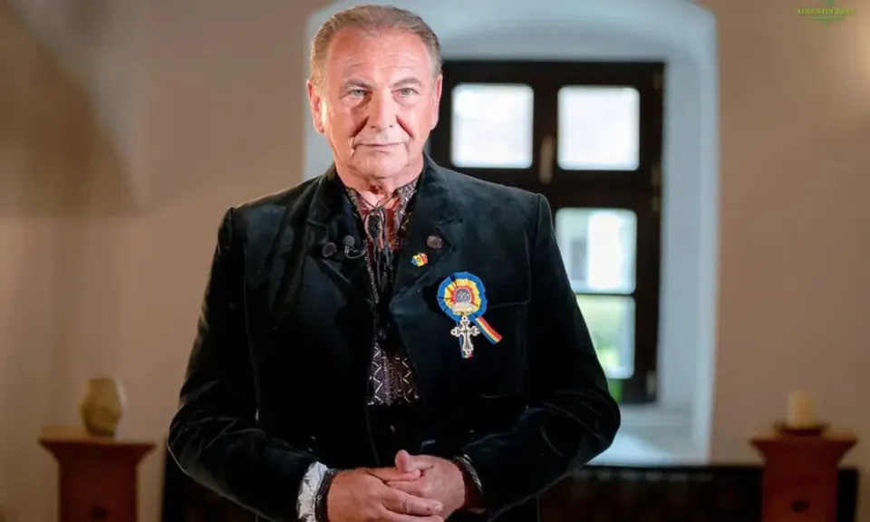 Un bărbat cu păr grizonat poartă o jachetă neagră, o cămașă cu model și o insignă colorată, în fața unei ferestre.