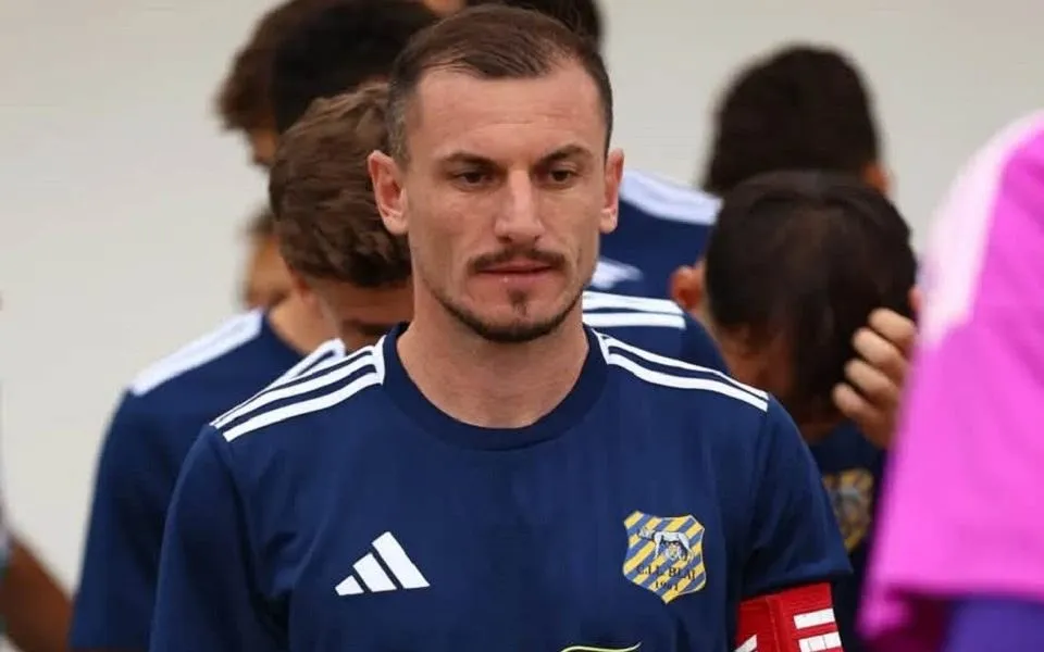 Un fotbalist în tricou bleumarin, cu banderolă roșie, stă în prim-plan, înconjurat de alți jucători și de o siluetă în roz.