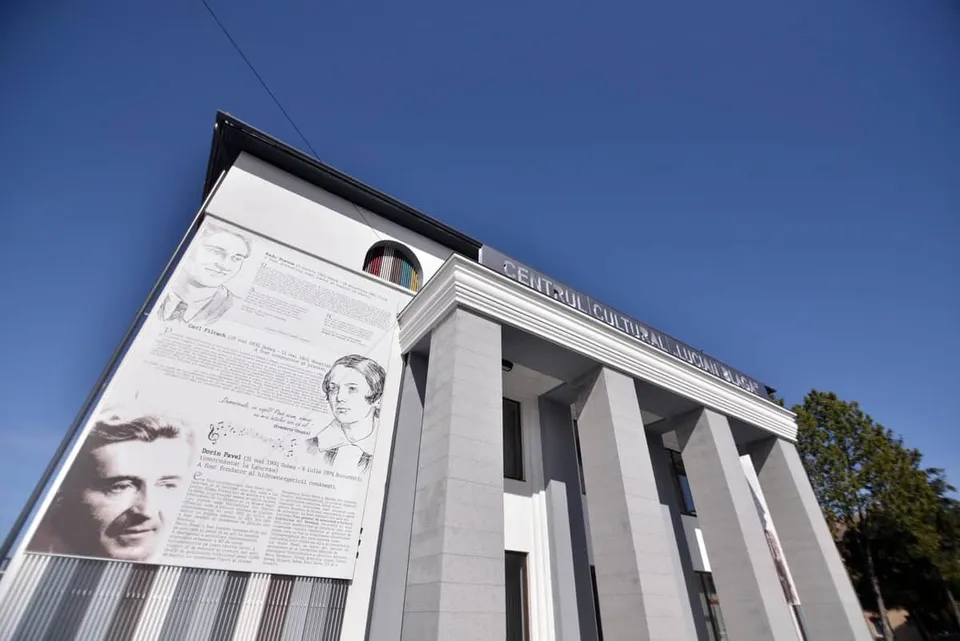 O clădire cu fațadă deschisă la culoare, coloane înalte și panouri cu portrete și text este fotografiată sub un cer albastru.