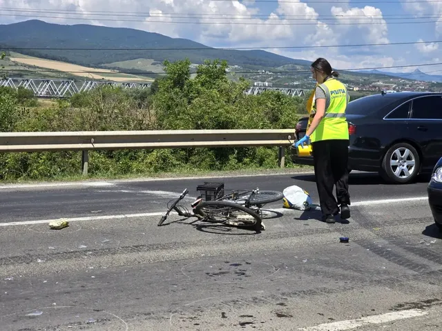 O bicicletă este căzută pe carosabil, lângă o cască albă; o persoană cu vestă reflectorizantă stă lângă un autoturism negru.