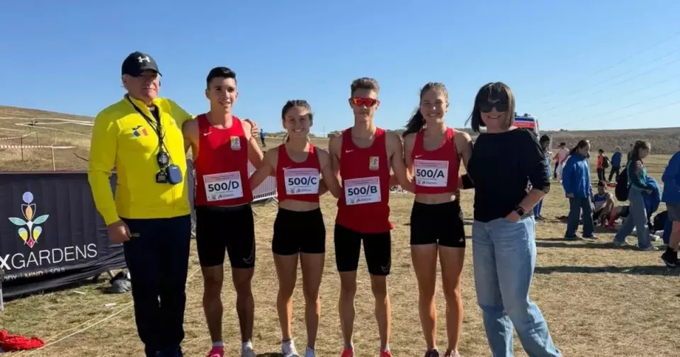 Șase persoane stau aliniate pe iarbă; patru poartă echipament sportiv cu numere de concurs, iar în fundal se văd alte persoane.