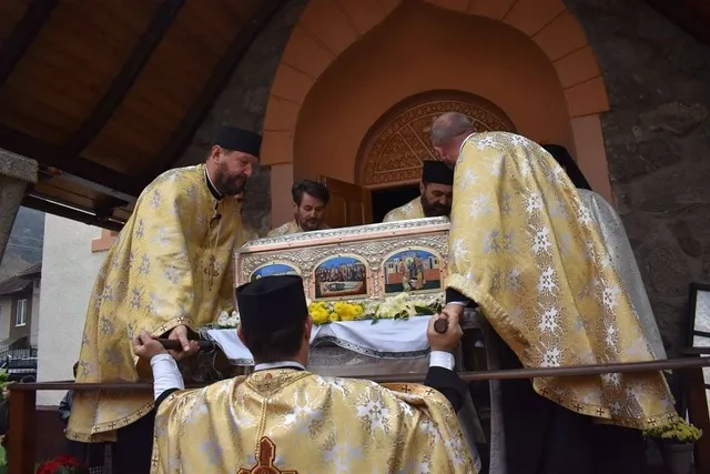 Patru bărbați în veșminte aurii stau în jurul unei racle ornamentate, așezate lângă flori galbene, sub o arcadă.