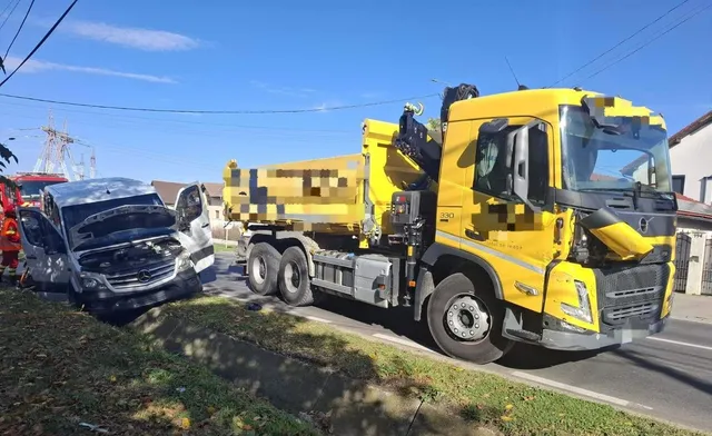 Un autoturism gri cu partea din față avariată se află lângă un camion galben, pe un drum, sub un cer senin.