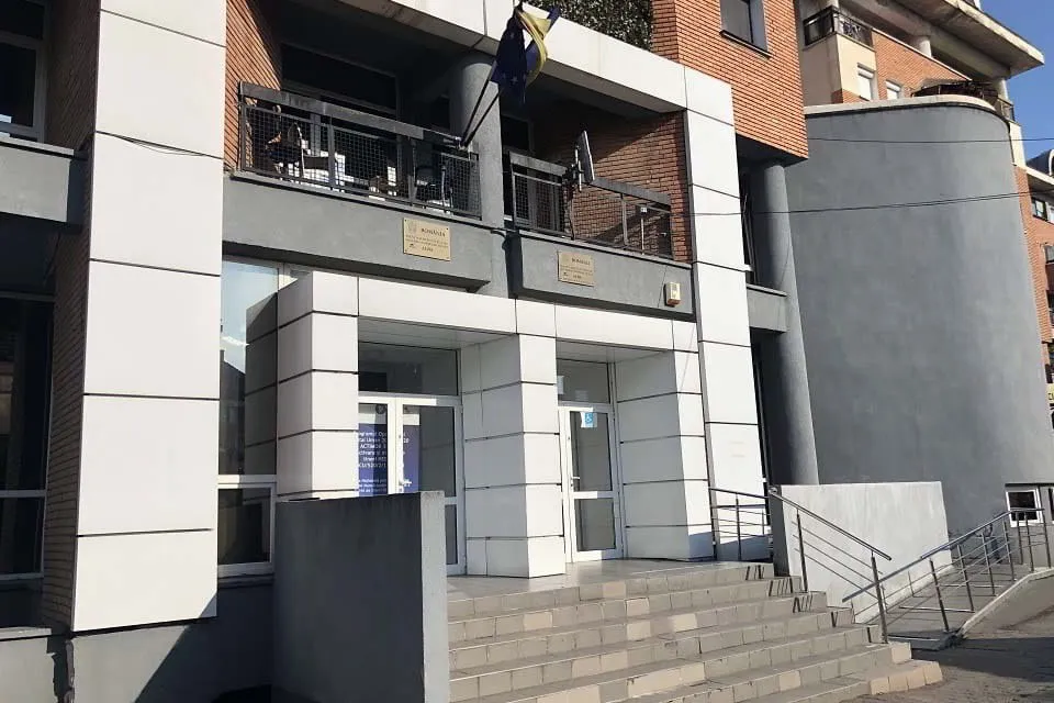 O clădire cu fațadă albă și maro are uși vitrate, balcoane, o structură cilindrică gri și trepte cu balustrade metalice.