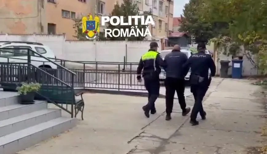 Doi polițiști în uniforme însoțesc un bărbat îmbrăcat în haine închise la culoare pe o alee, lângă trepte și o balustradă.