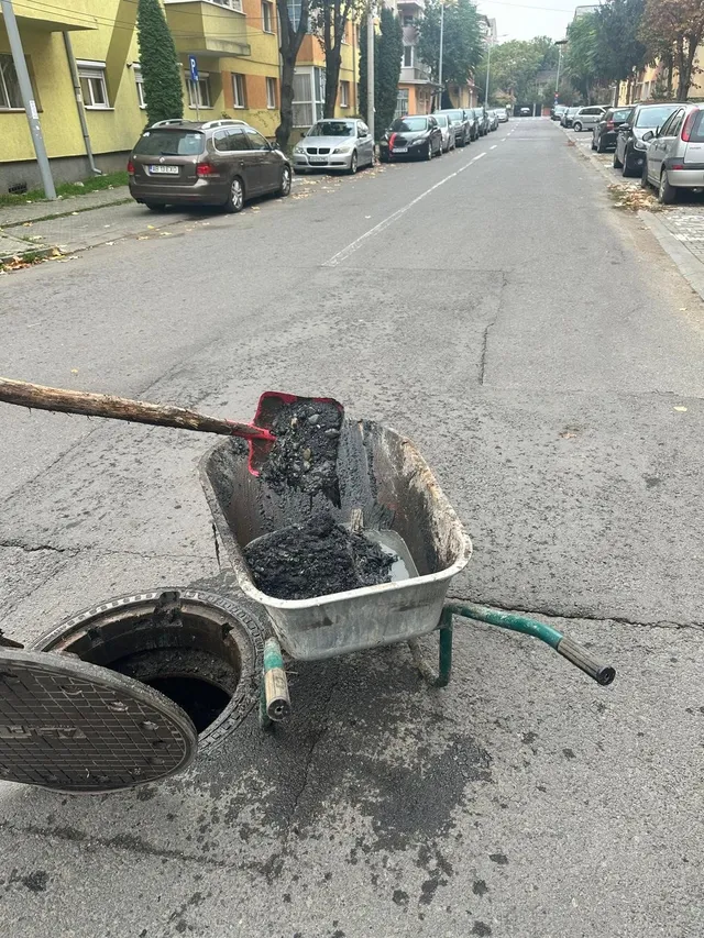 În prim-plan, un capac de canal ridicat stă lângă o gură de canal deschisă și o roabă cu material negru; mașini sunt parcate de-a lungul străzii.