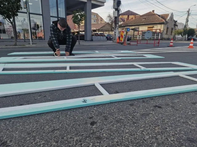 O persoană îmbrăcată într-o jachetă în carouri și pantaloni închiși la culoare stă aplecată lângă marcaje albe și turcoaz pe asfalt.