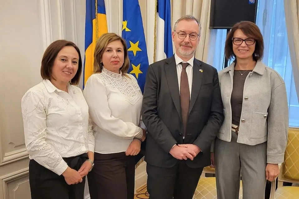 Patru adulți stau unul lângă altul într-o încăpere, în fața unor steaguri; două femei poartă bluze albe, iar celelalte persoane poartă sacouri.