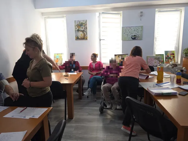 Mai multe persoane stau la mese într-o cameră luminoasă; o persoană stă într-un scaun cu rotile, iar pe mese sunt hârtii.