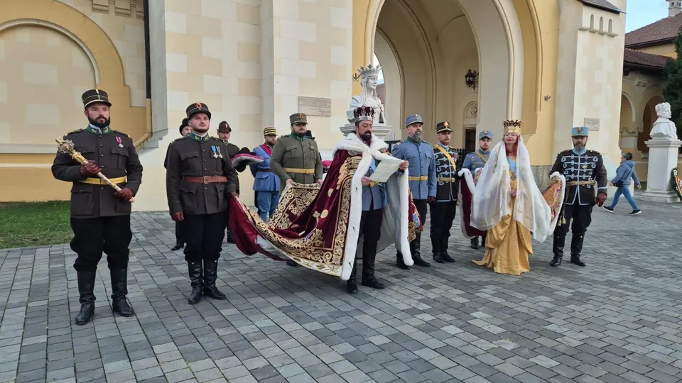 Mai multe persoane în costume de epocă stau în fața unei clădiri cu arcade; două poartă mantii lungi și coroane.