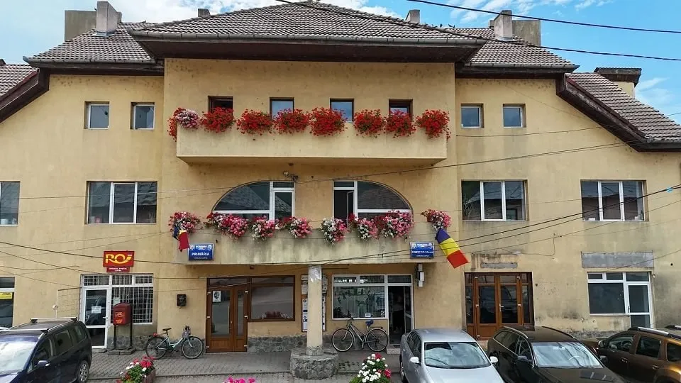 O clădire deschisă la culoare are balcoane cu flori și steaguri; în față sunt parcate mașini și biciclete.