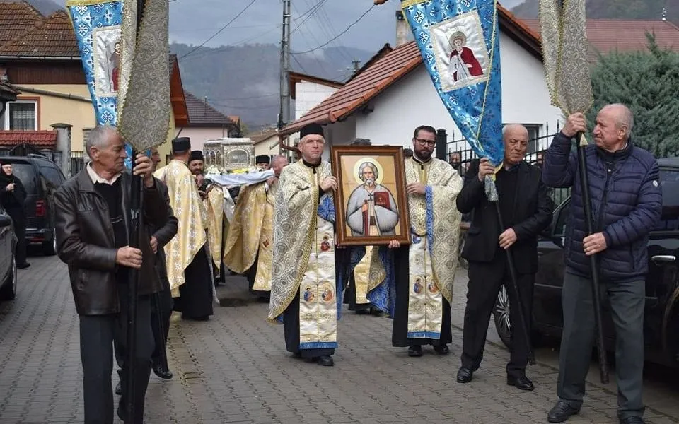 Mai mulți bărbați stau pe o stradă; unii poartă veșminte deschise la culoare, iar alții țin prăjini cu pânze albastre.