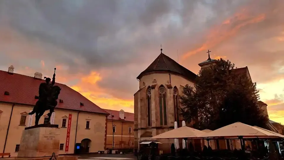 O clădire cu acoperiș înalt și ferestre înguste se află lângă alte clădiri; în față se văd un monument ecvestru și umbrele deschise.