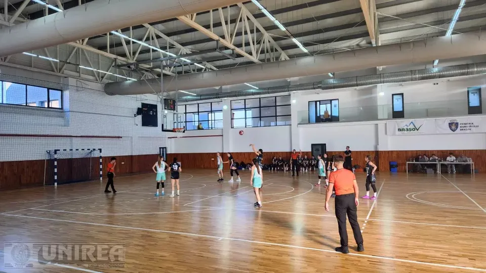 Într-o sală de sport, jucătoare în echipament sportiv se află pe terenul de baschet, iar o persoană în vestă portocalie stă în prim-plan.