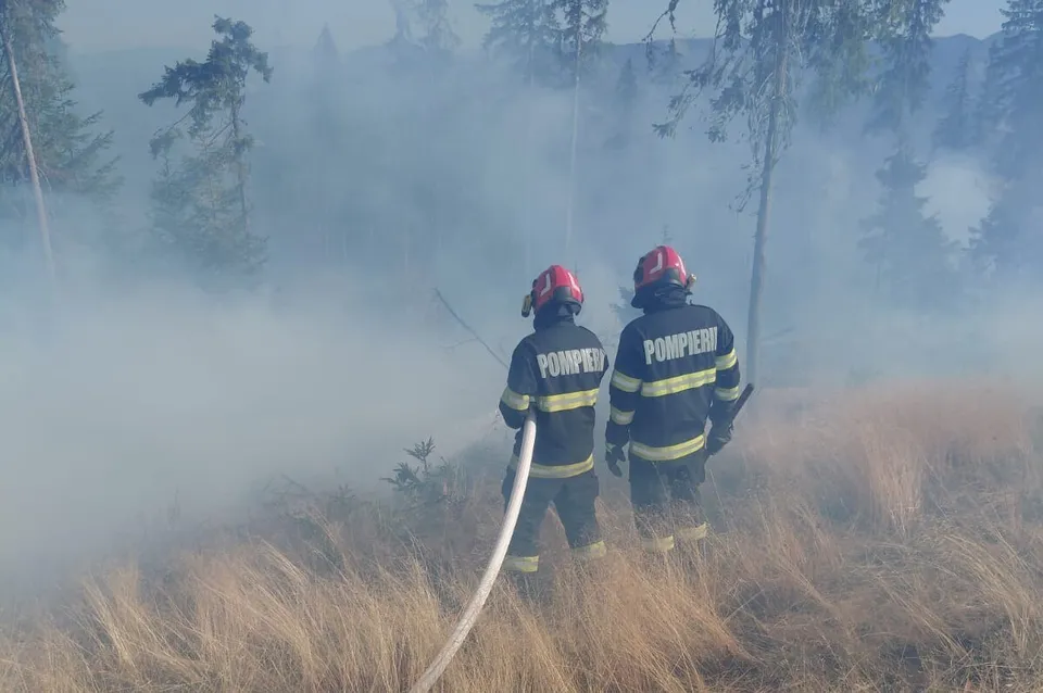 Doi pompieri cu căști roșii și uniforme bleumarin stau în vegetație uscată, ținând un furtun alb, în mijlocul fumului dens.