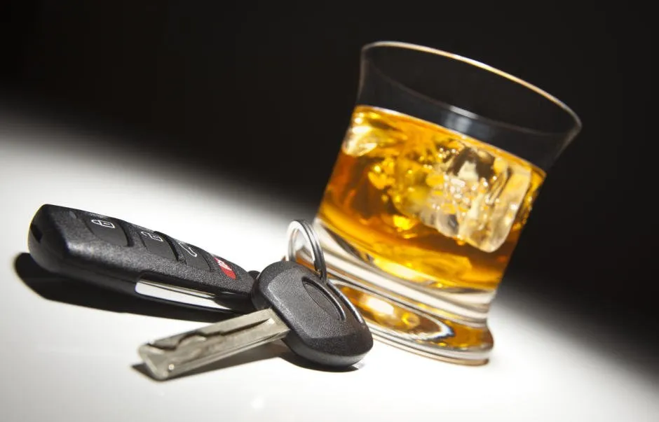 Un pahar scund cu lichid auriu și cuburi de gheață stă lângă două chei auto, pe o suprafață albă.