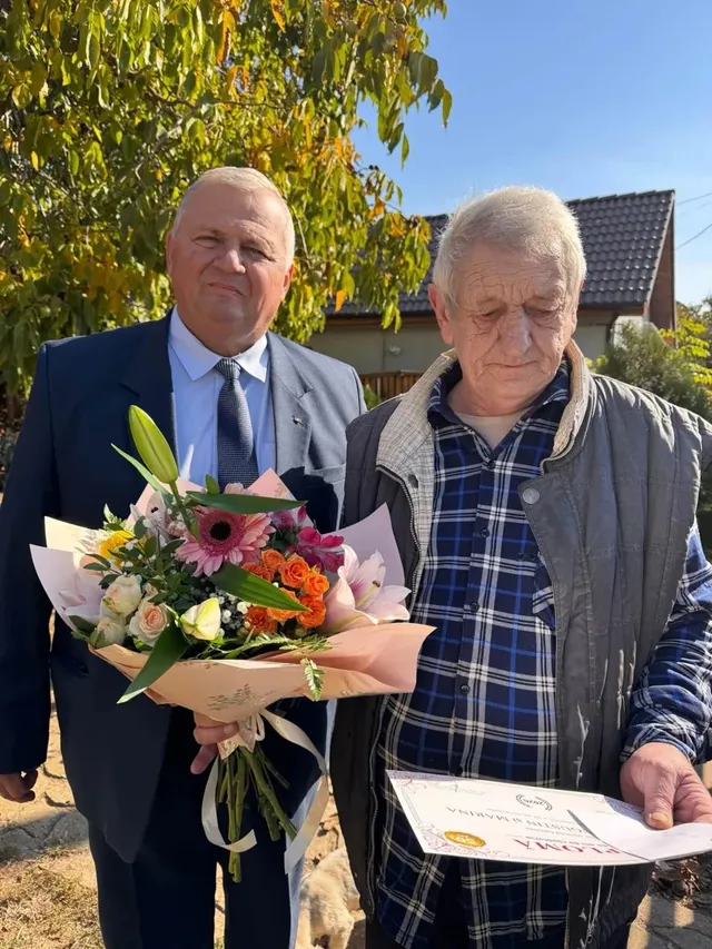 Un bărbat în costum stă lângă un bărbat în vârstă, care ține un buchet de flori și o diplomă.