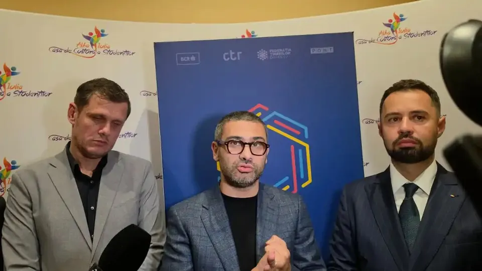 Trei bărbați în sacouri stau în fața unui panou cu sigle; bărbatul din mijloc poartă ochelari și ține mâinile ridicate.