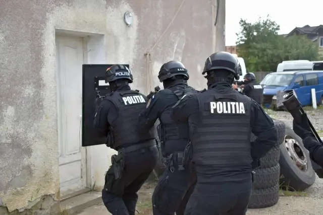Mai mulți polițiști în uniforme și veste inscripționate stau lângă o ușă, iar unul ține un scut negru.