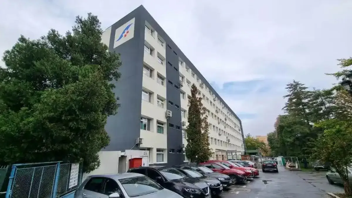 O clădire înaltă, albă și gri, cu multe ferestre, se află lângă un șir de mașini parcate și copaci.