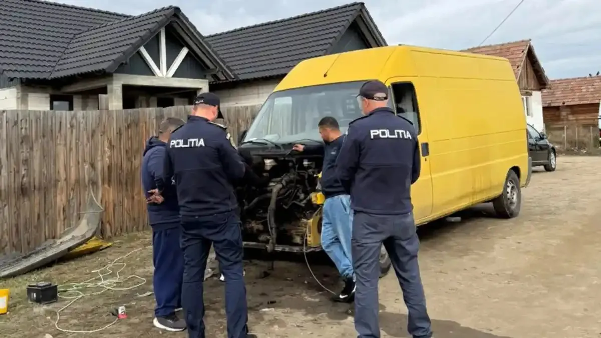 Trei persoane în uniforme bleumarin stau lângă partea din față desfăcută a unei autoutilitare galbene, pe un drum neasfaltat.