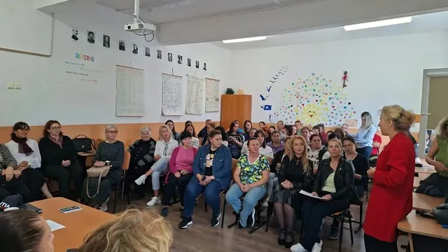 Mai multe persoane stau pe scaune într-o sală, iar o femeie îmbrăcată în roșu stă în picioare lângă mese.