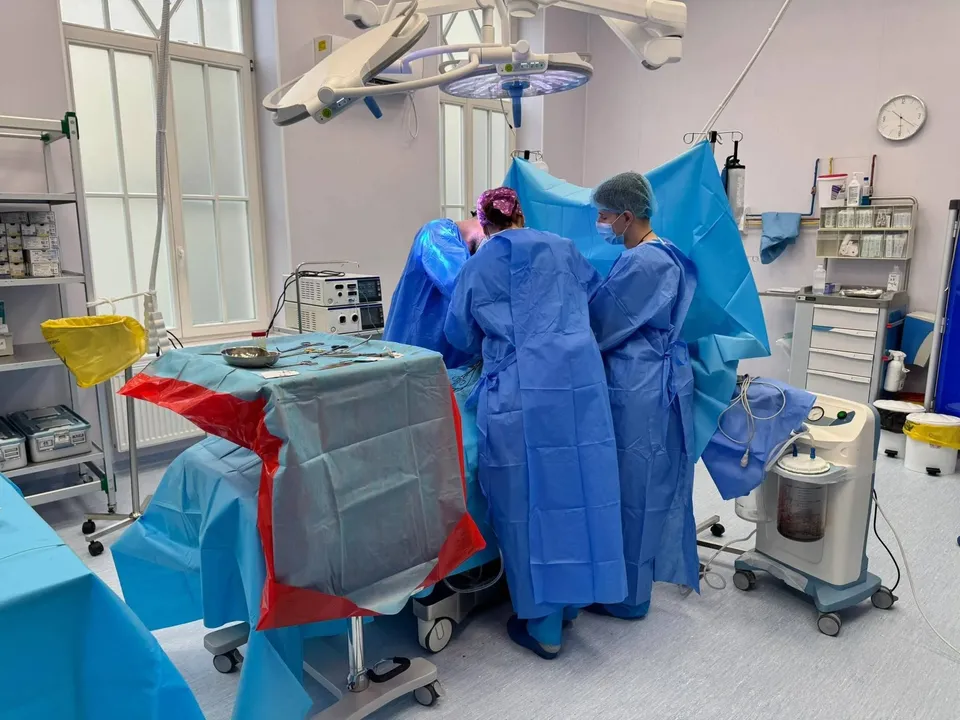 Două persoane în halate albastre, cu măști și bonete, stau lângă o masă acoperită cu material albastru și roșu, între aparate medicale.