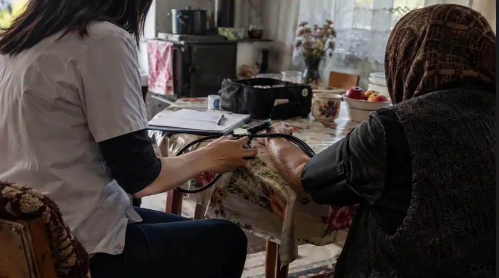 Două persoane stau la o masă; una poartă o bluză deschisă la culoare și ține un obiect lângă brațul întins al celeilalte.