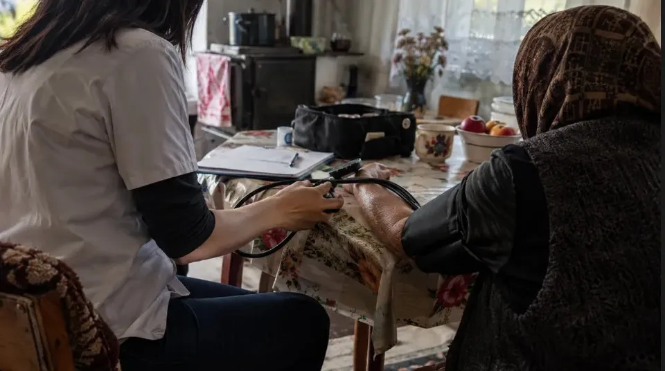 Două persoane stau la o masă; una poartă o bluză deschisă la culoare și ține un obiect lângă brațul întins al celeilalte.