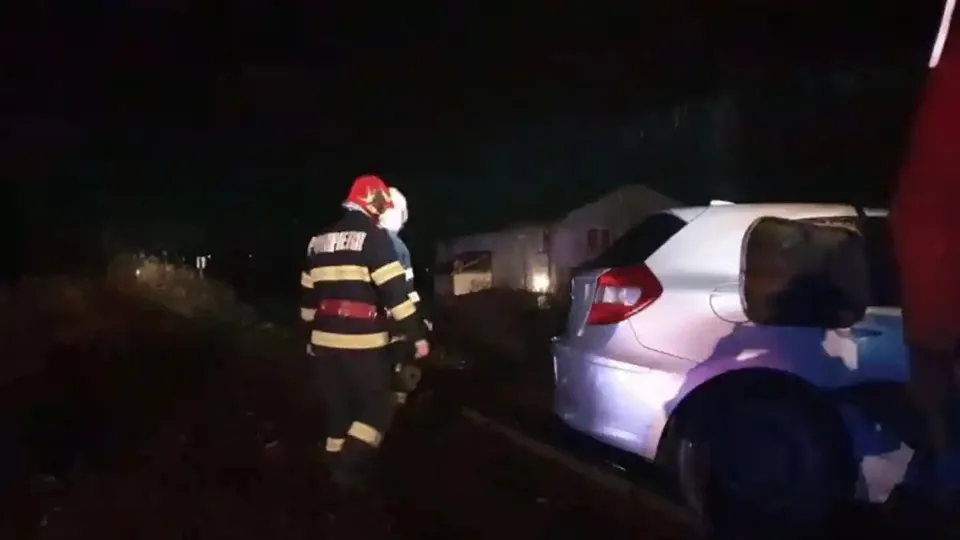 O persoană cu cască roșie și echipament cu benzi reflectorizante stă lângă un autoturism argintiu, într-un cadru întunecat.